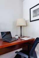 Un escritorio de madera con un ordenador portátil, una lámpara, una pequeña planta en maceta y un cuadro en la pared.