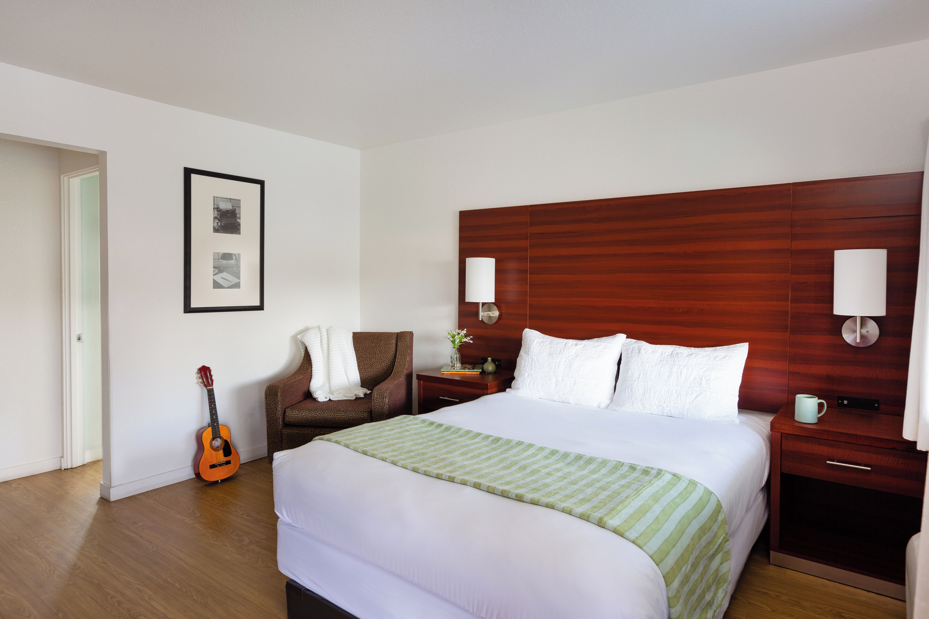 Habitación de hotel con una cama grande, cabecero de madera, una guitarra, una silla y un cuadro en la pared.