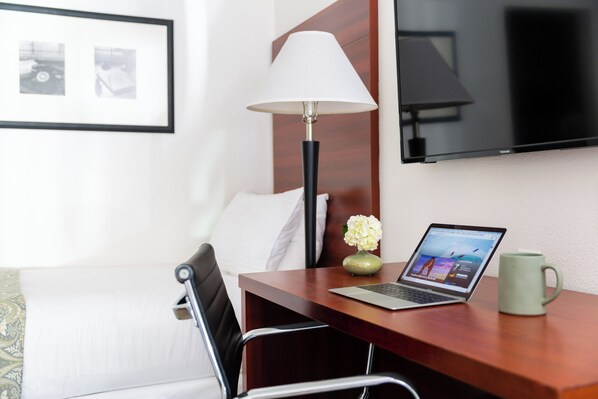 Une chambre d’hôtel avec un lit, un bureau avec un ordinateur portable, une lampe, un vase de fleurs et un téléviseur fixé au mur.