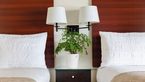 Une chambre d’hôtel avec deux lits, du linge de lit blanc, une table de chevet ornée d’une plante en pot et deux lampes fixées au mur.
