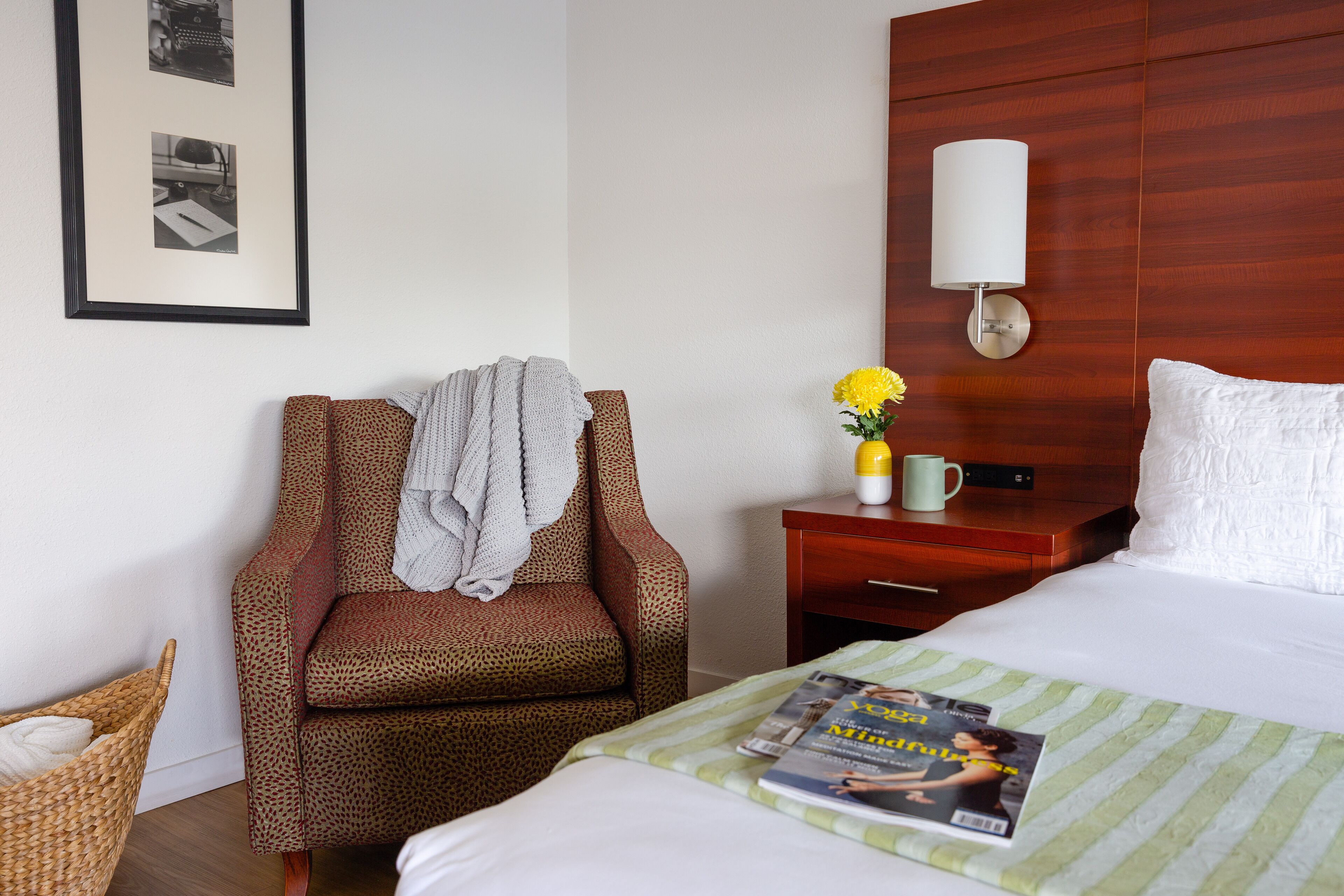 A hotel room with a bed, a chair with a blanket, a nightstand with a lamp, a vase with flowers, and a framed picture on the wall.
