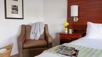 A hotel room with a bed, a chair with a blanket, a nightstand with a lamp, a vase with flowers, and a framed picture on the wall.
