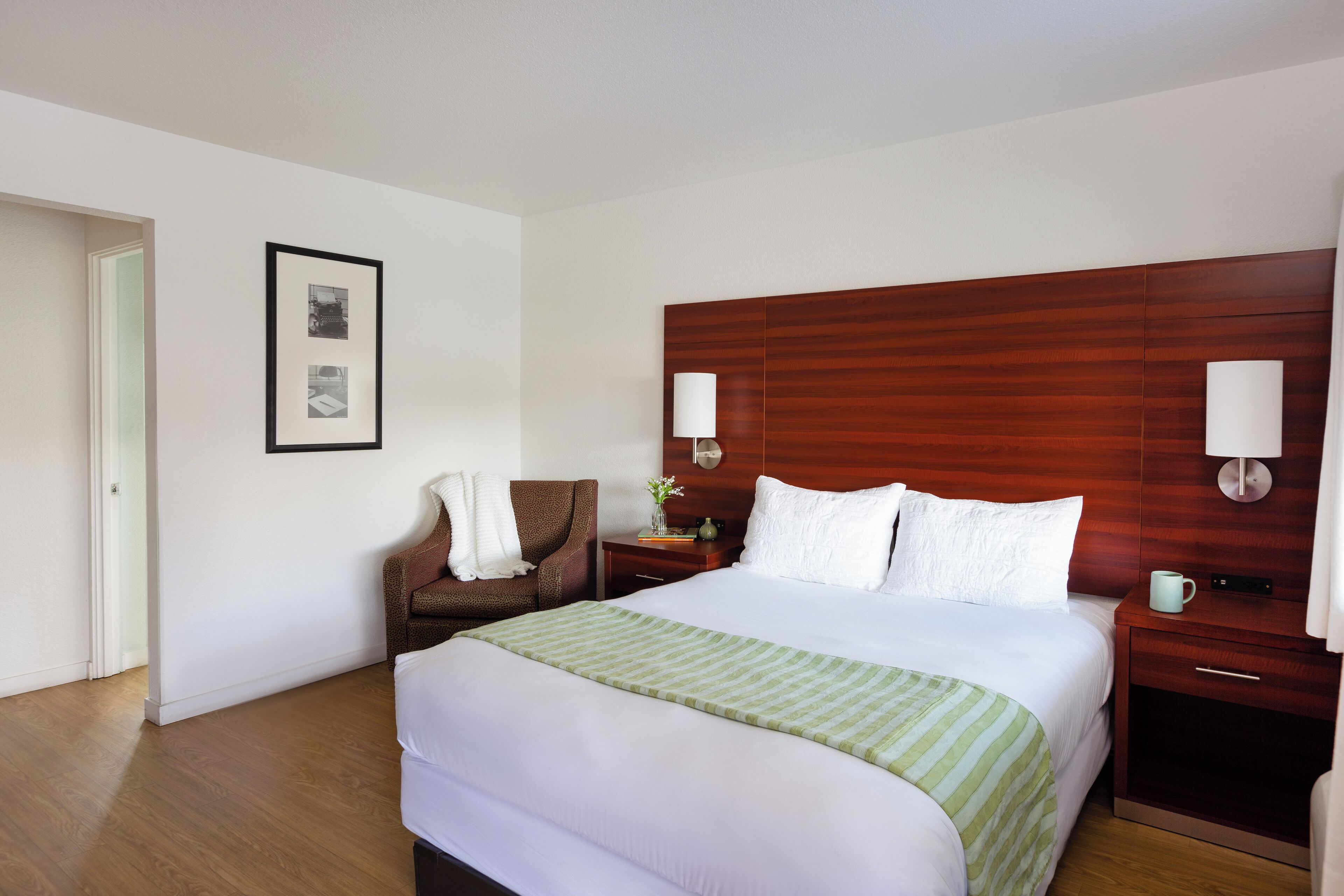 Une chambre d’hôtel avec un grand lit, une tête de lit en bois, deux lampes de chevet, une chaise, une table de chevet et un tableau encadré au mur.
