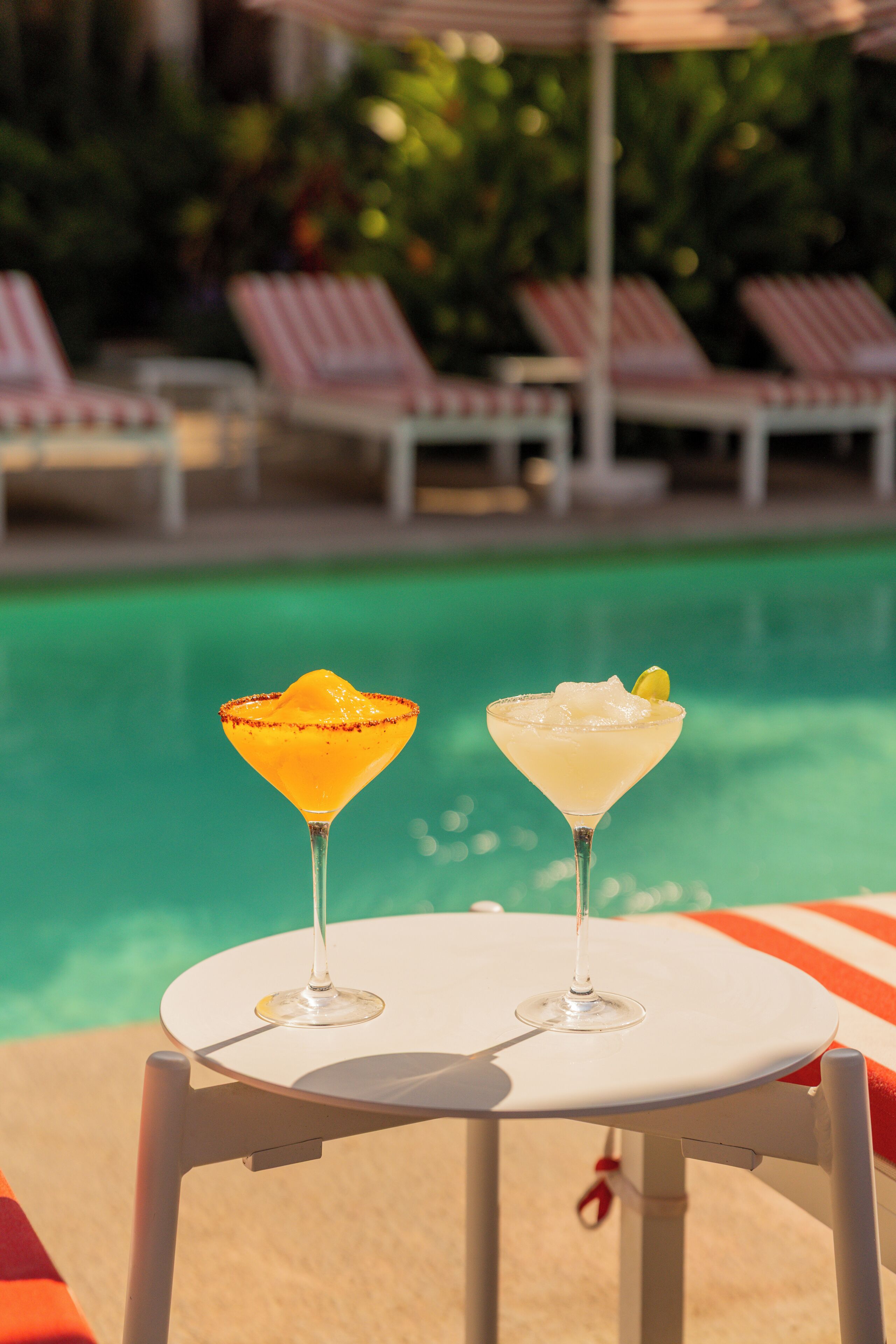 Two cocktails on a white table by a poolside with lounge chairs in the background.