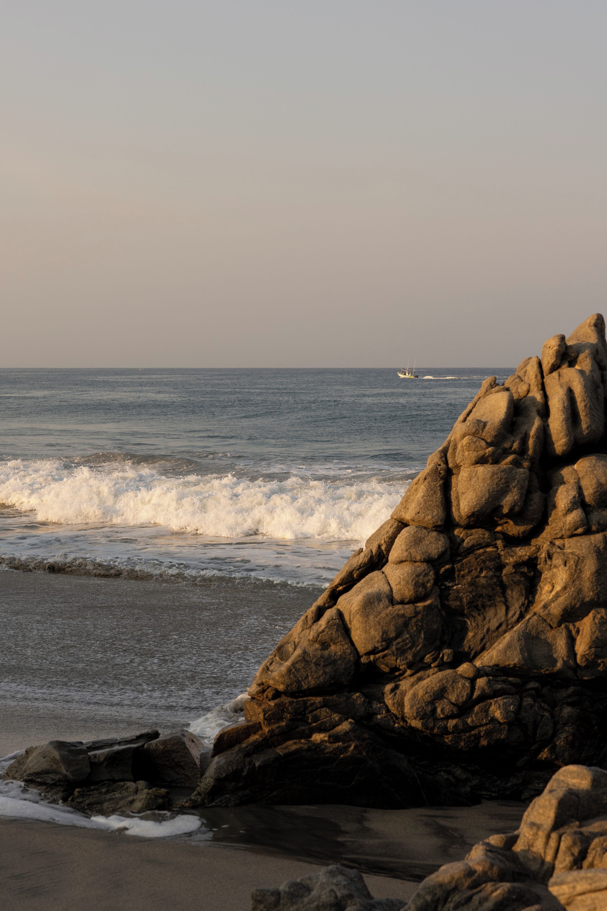 A rocky shoreline with waves, a boat in the distance, and a clear sky.