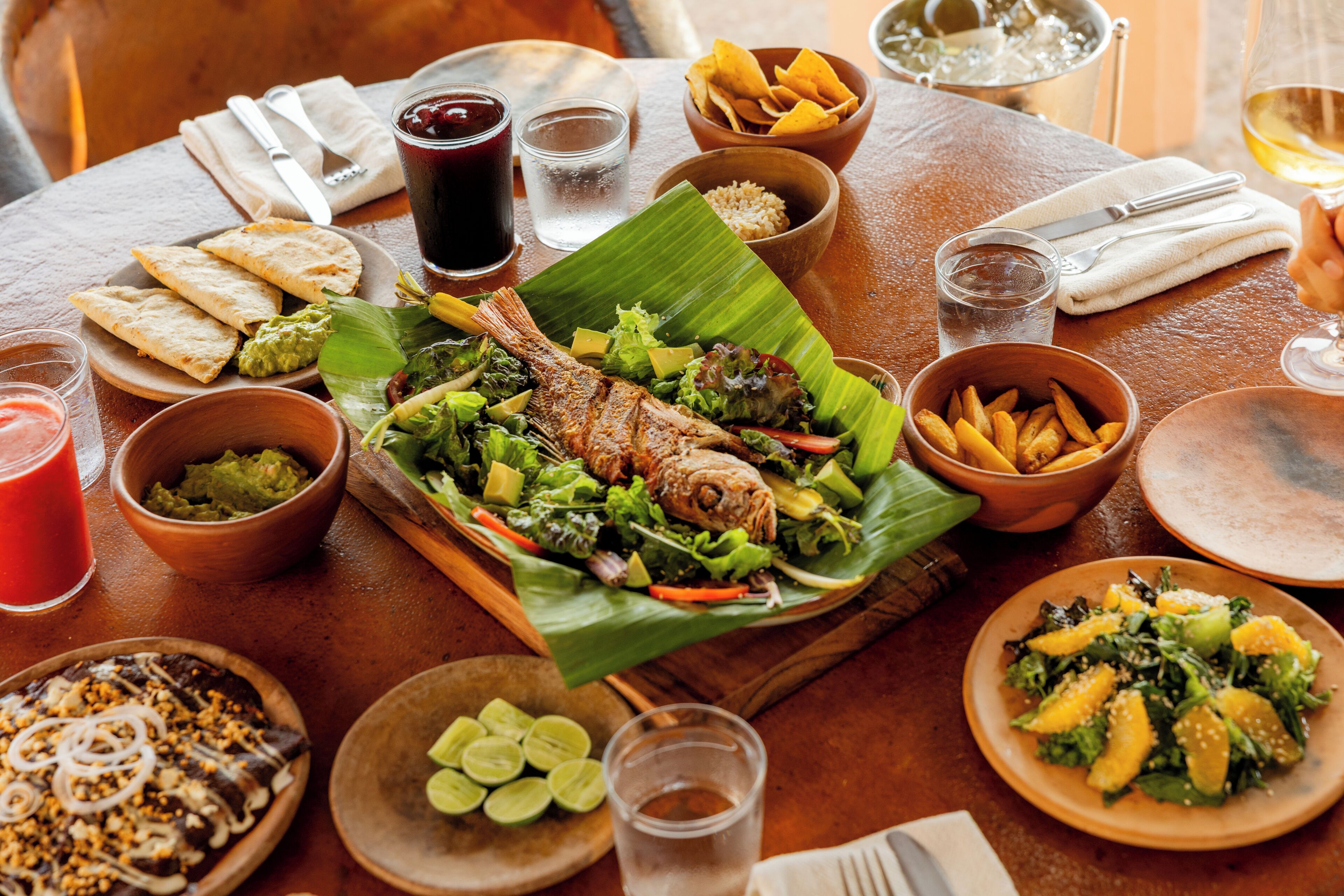 Una mesa de comedor con diversos platos, incluyendo un pescado sobre una hoja de plátano, ensaladas y pan.