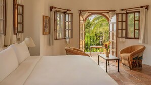 Habitación de hotel con una cama, dos sillas de mimbre, una mesita con un florero y un balcón con vistas a zonas verdes.