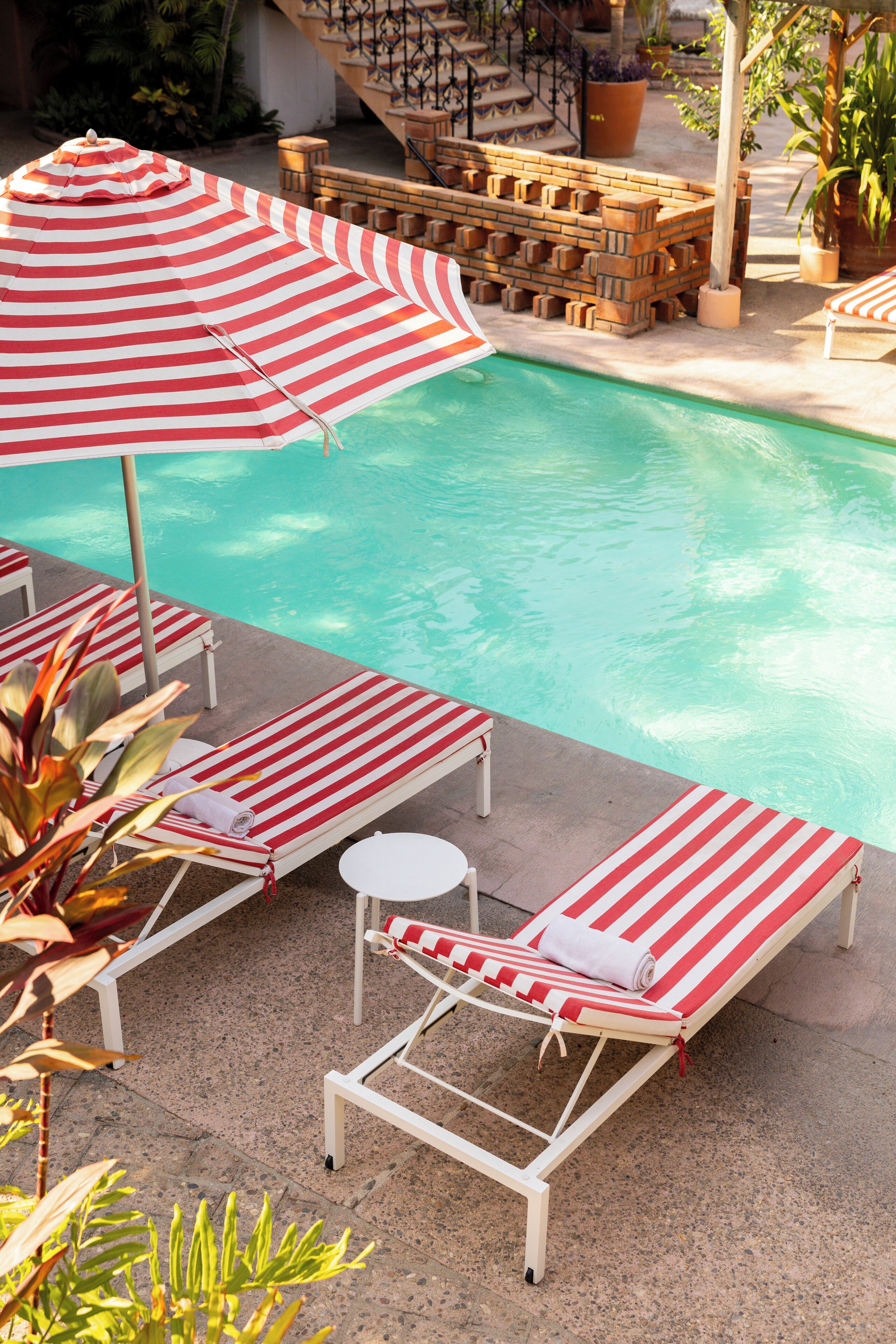 Un espace aménagé au bord de la piscine, avec des chaises longues à rayures et des parasols, un mur en briques et des plantes en pot.