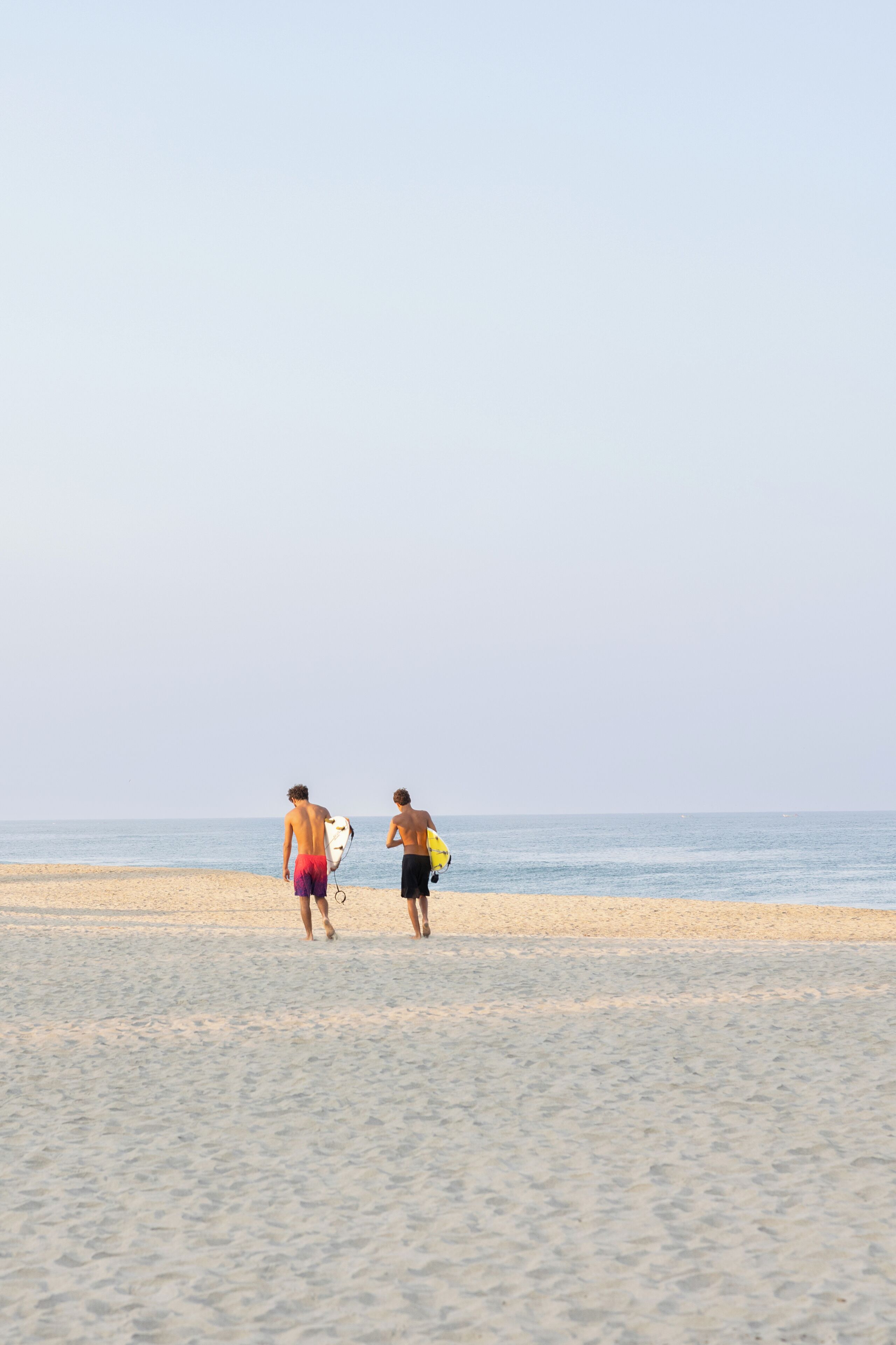 Twee mensen wandelen over een zandstrand, met een rustige zee op de achtergrond.