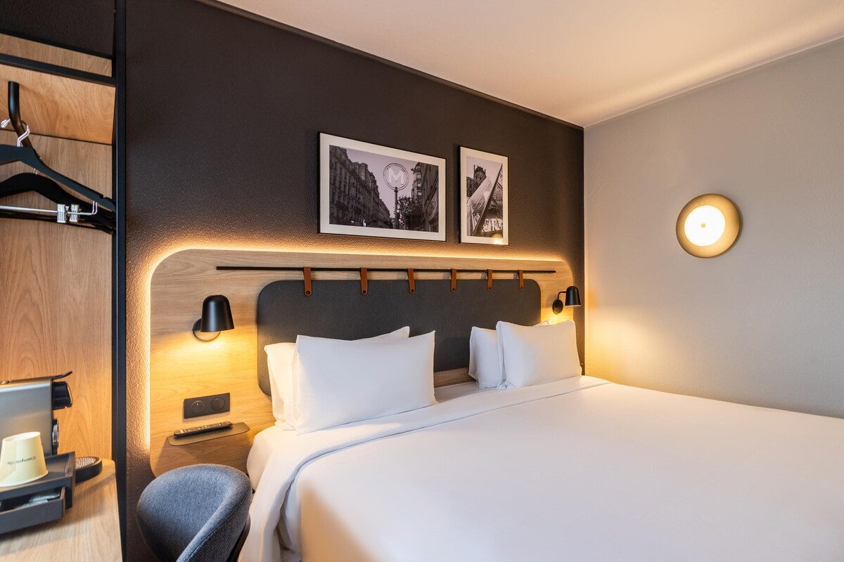 Een hotelkamer met een groot bed, een nachtkastje met een lamp, een stoel, een kapstok en twee ingelijste foto's aan de muur.