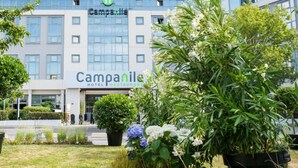 Un hotel moderno con un llamativo letrero de 'Campanile', que presenta fachada de vidrio y un jardín ajardinado con diversas plantas y flores.