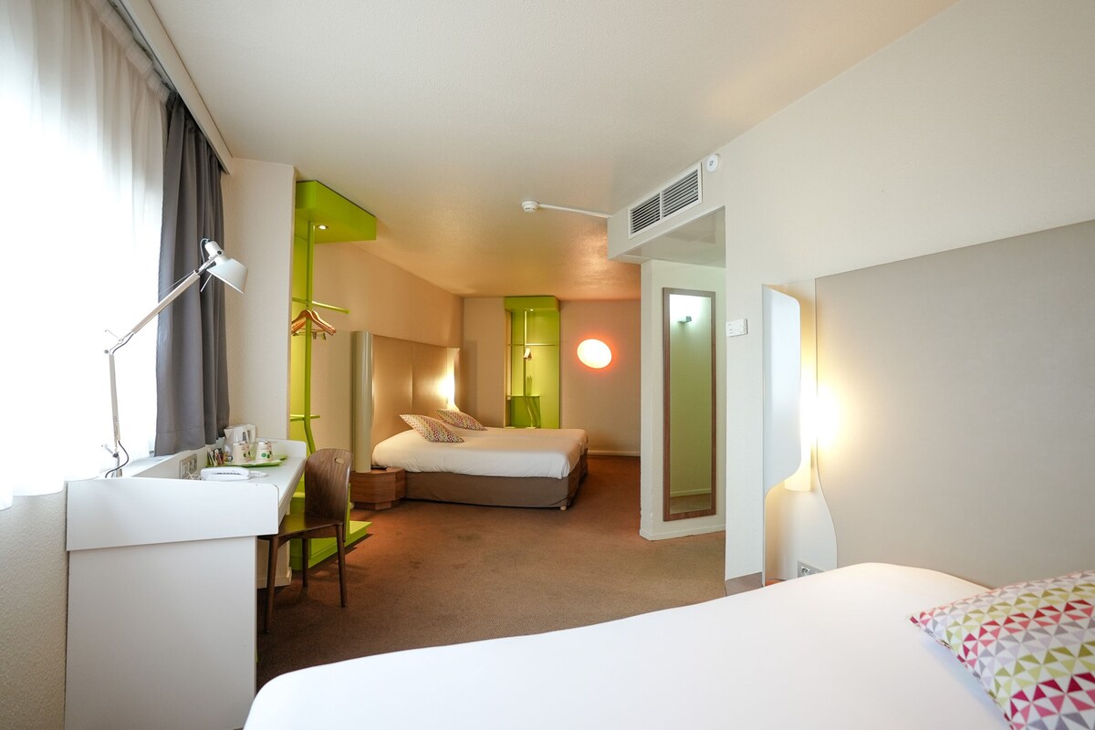 A hotel room with a bed, a desk, a chair, and a bedside table.