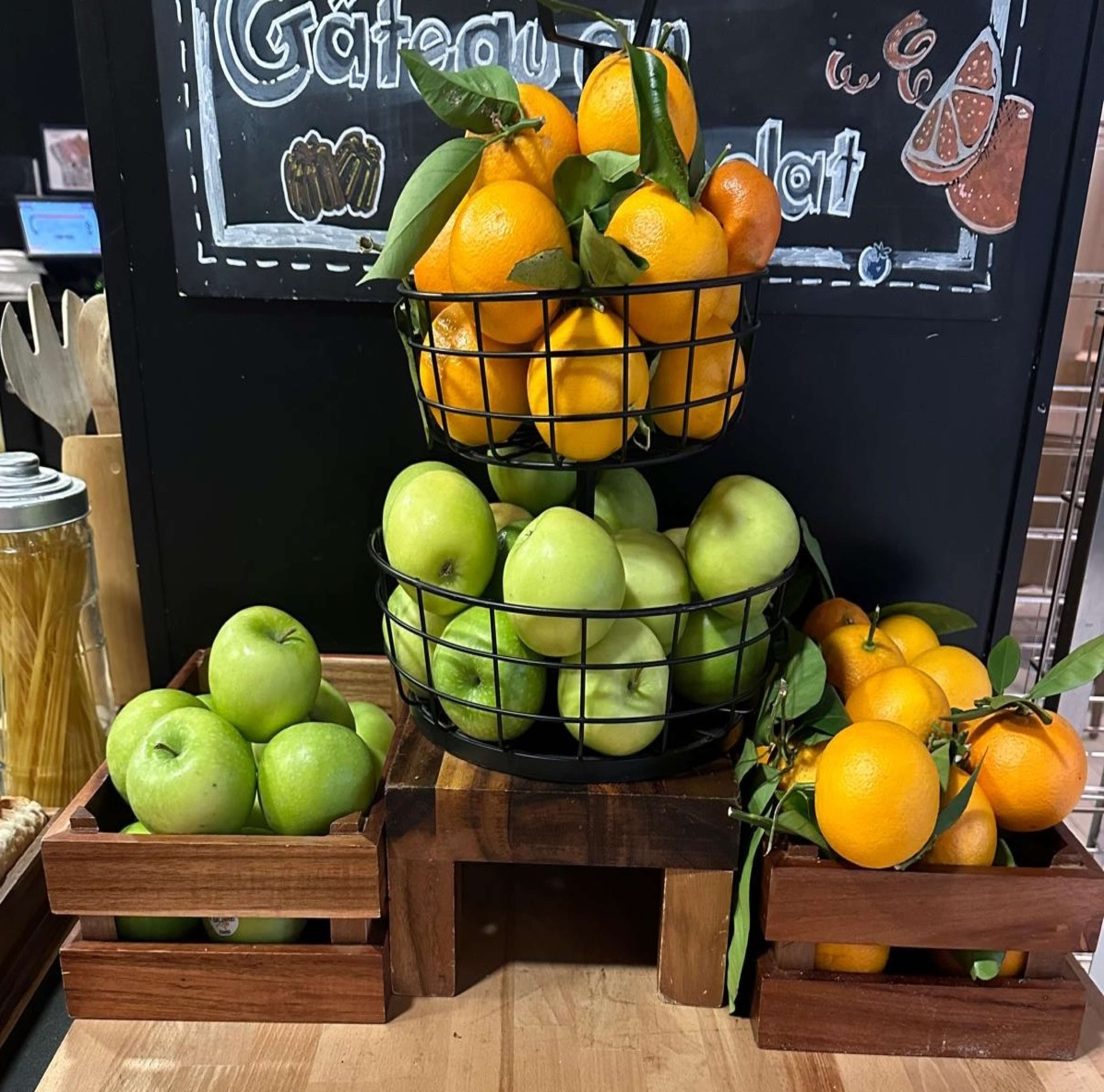 Una exhibición de frutas en cestas, con manzanas verdes y naranjas, y un pizarrón al fondo.