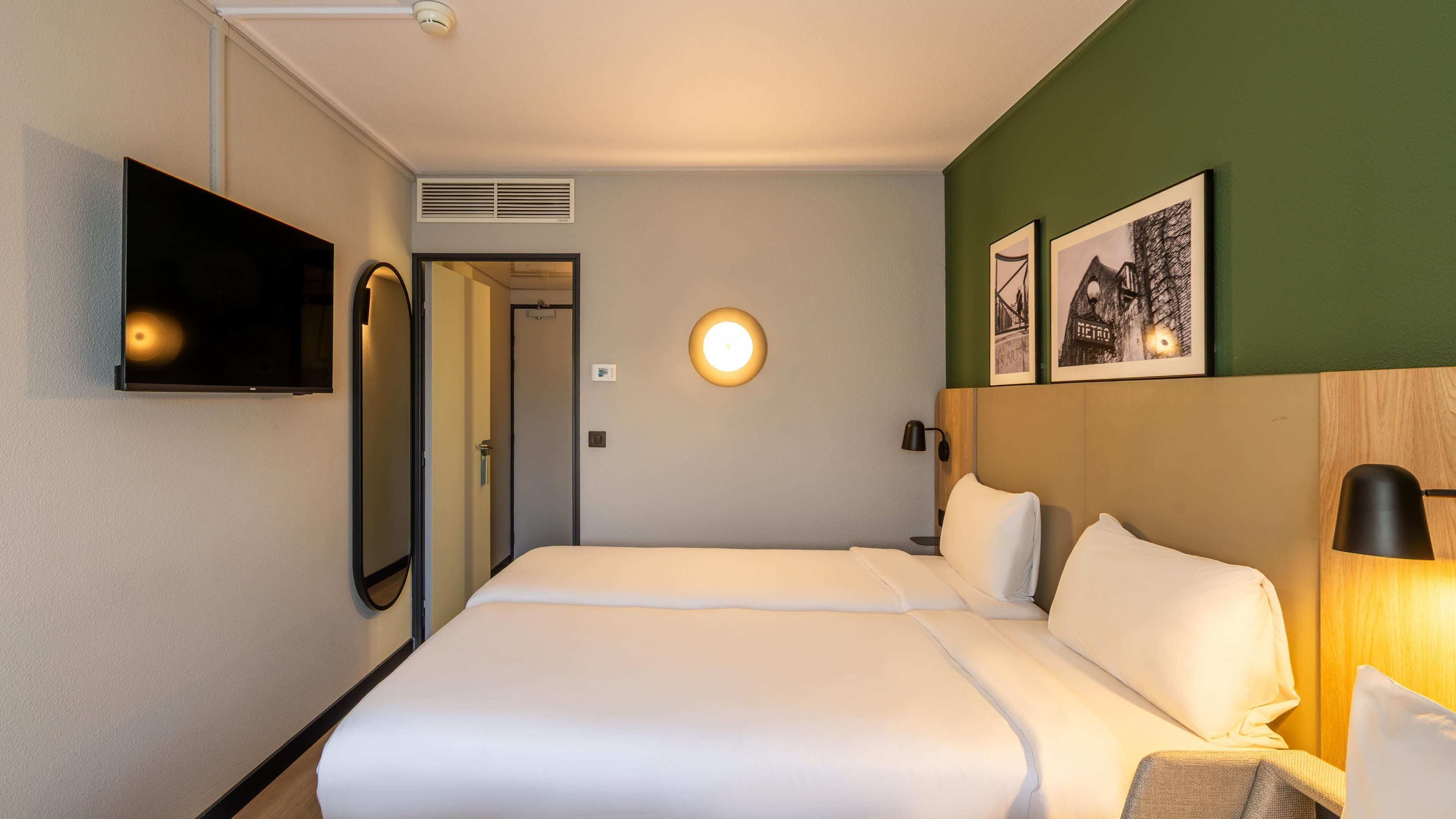 A hotel room with a bed, a TV mounted on the wall, a round wall light, and two framed pictures.