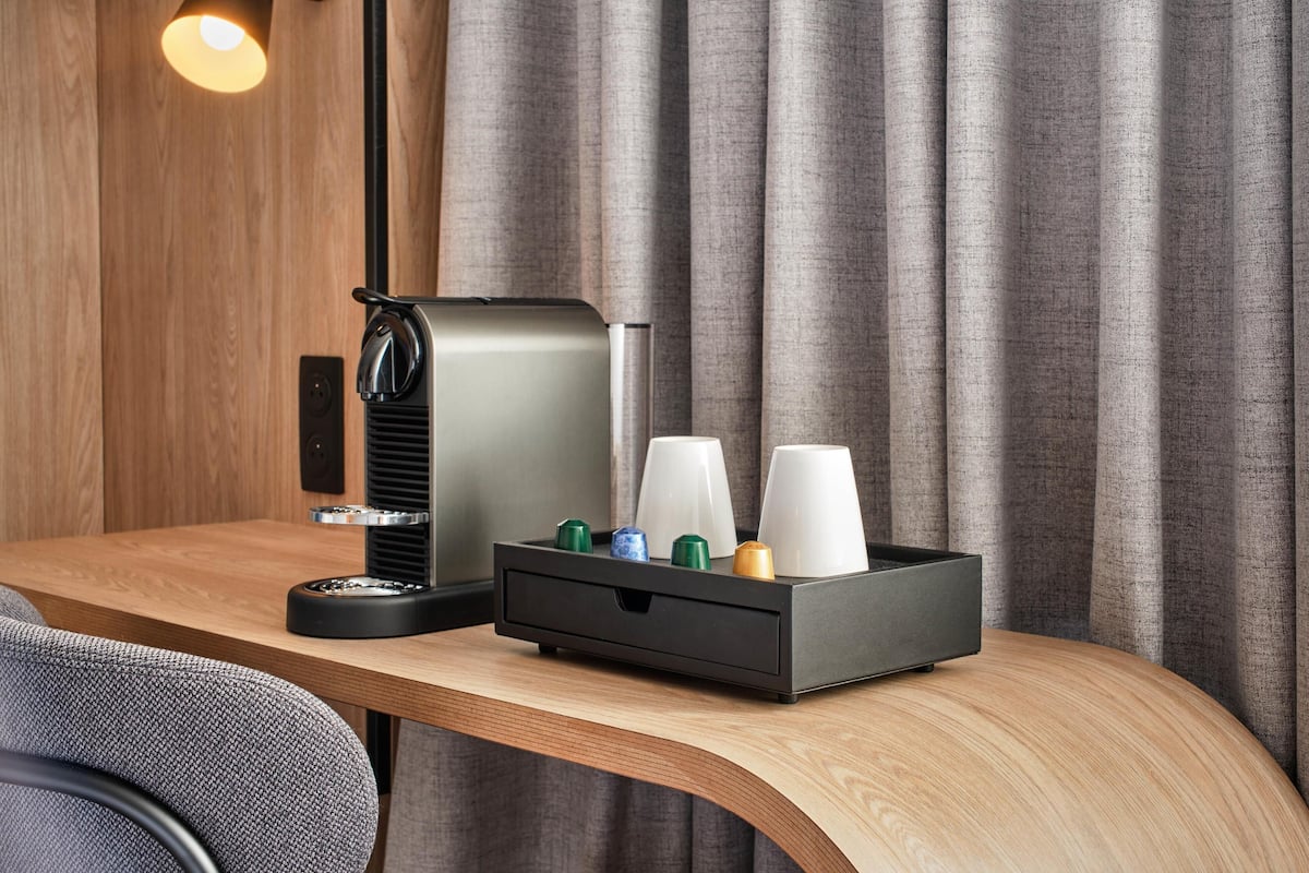 Une chambre d’hôtel moderne avec un bureau en bois, une machine à café noire et un plateau avec des tasses et une bouteille.