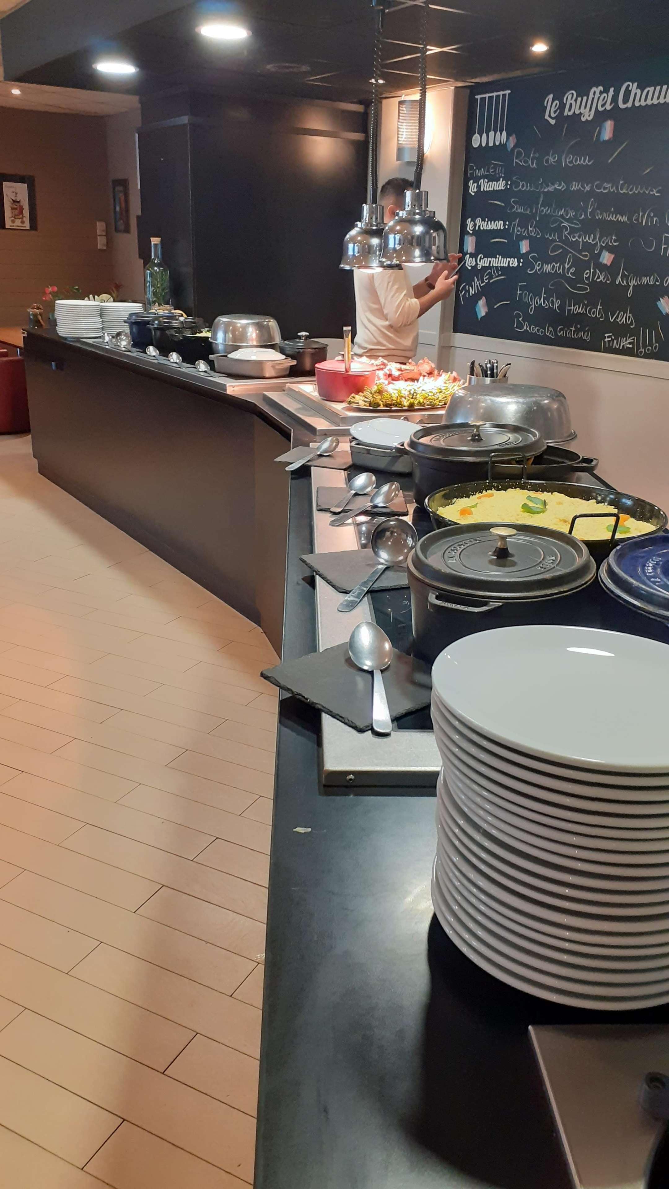 Zona de bufé con diversos alimentos, utensilios, un menú en pizarra y una persona sirviendo.
