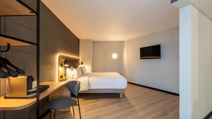 Habitación de hotel con cama, escritorio y silla, televisor montado en la pared y lámpara redonda.