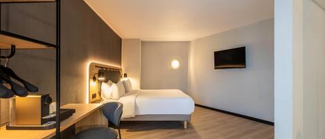 A hotel room with a bed, a desk with a chair, a TV mounted on the wall, and a round light fixture.