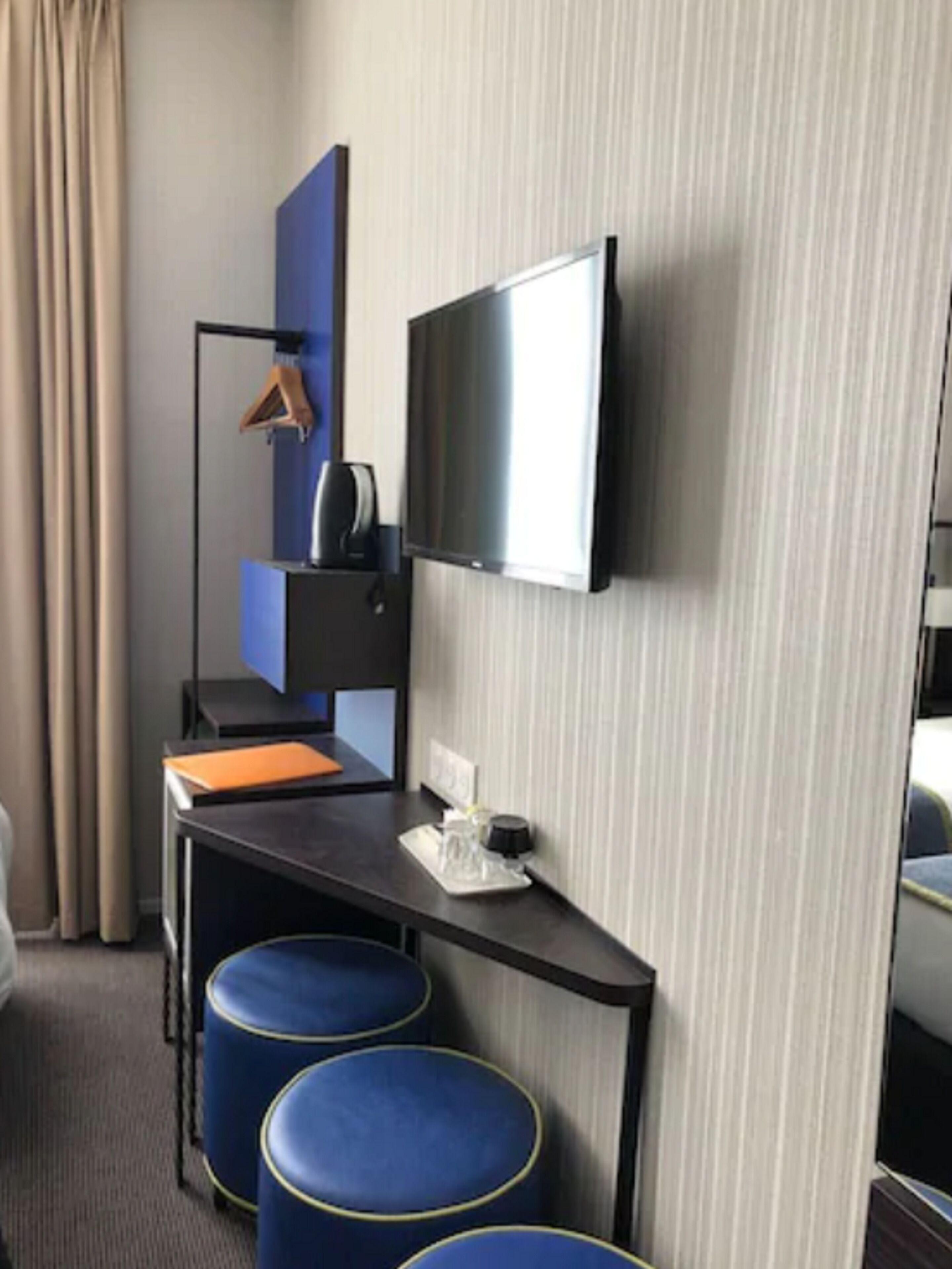 A hotel room with a flat-screen TV mounted on the wall, a desk with a chair, a small table with a cup and a tissue box, and two blue ottomans.