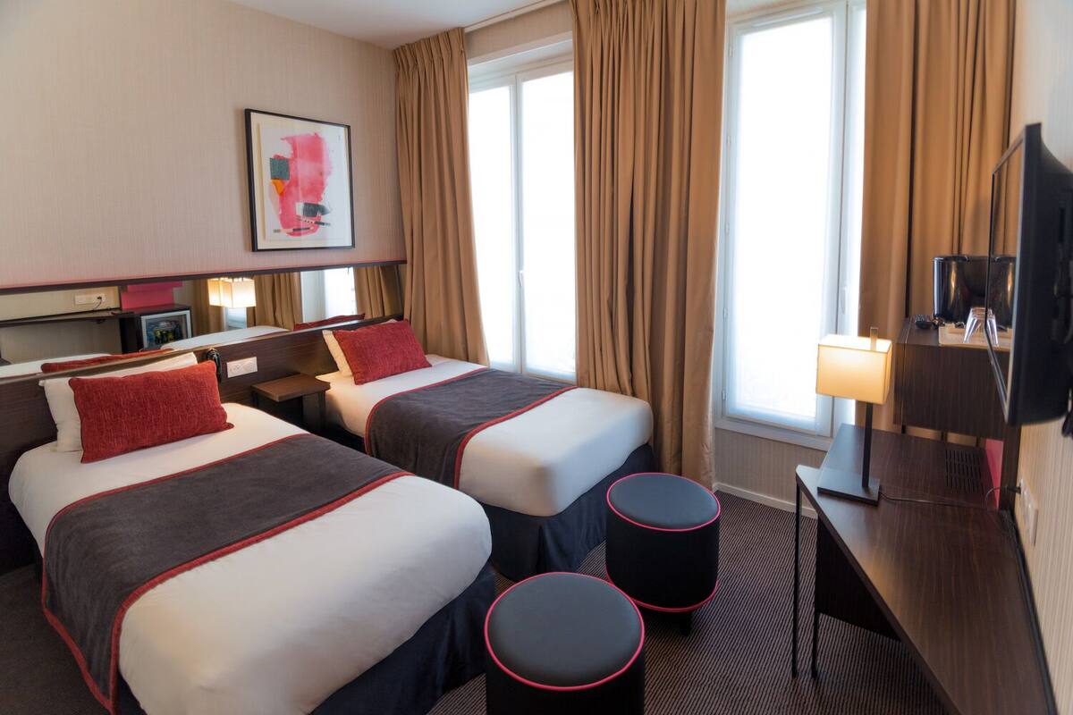Ein Hotelzimmer mit zwei Betten, einem Schreibtisch, einem Fernseher und einem Fenster mit Vorhängen.