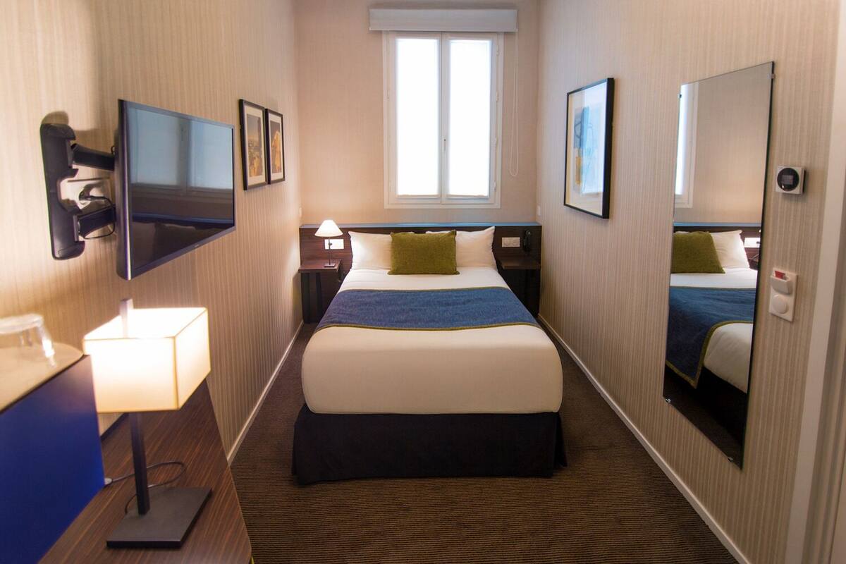 Ein Hotelzimmer mit Bett, Wandfernseher, Nachttischlampe, Fenster und Spiegel.