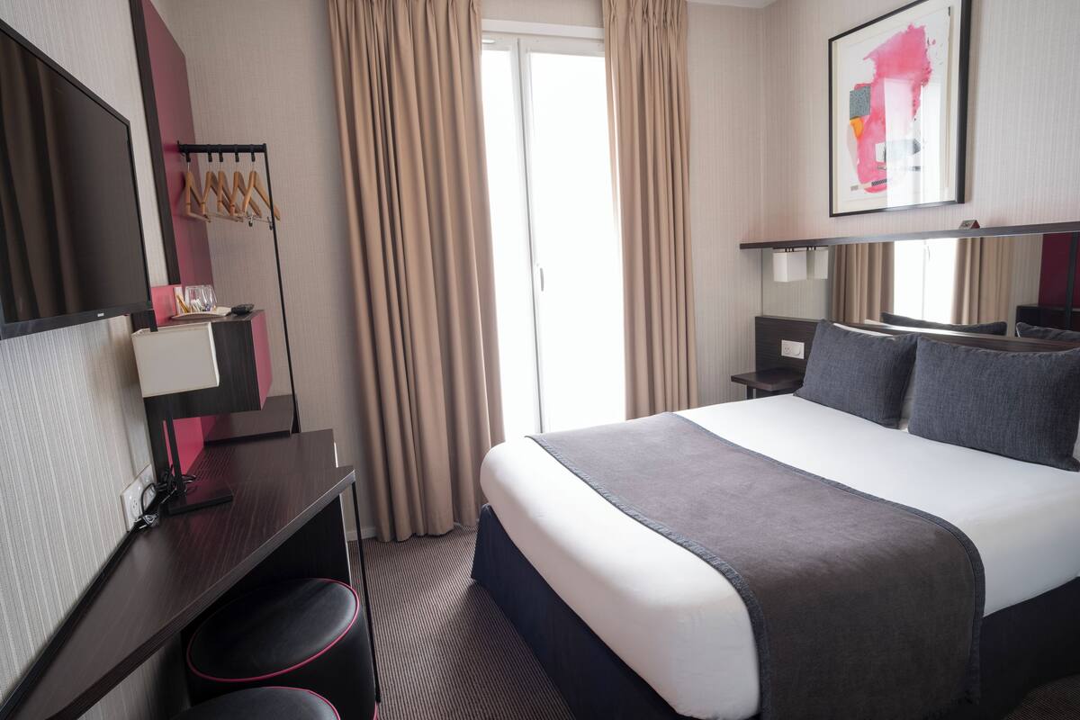 Ein Hotelzimmer mit einem großen Bett, einem Schreibtisch mit Stuhl, einem Fernseher und einem Fenster mit Vorhängen.