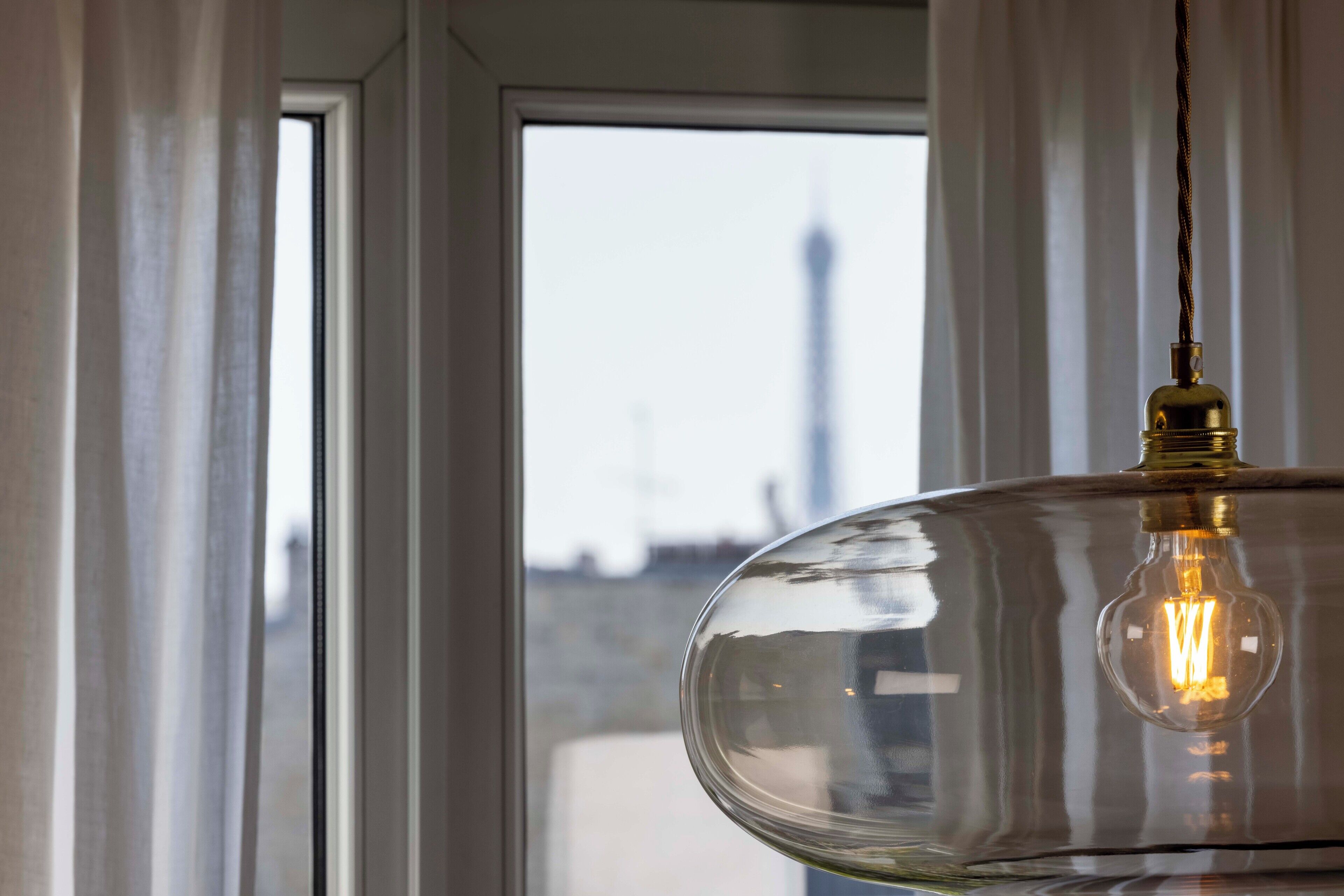 A clear glass pendant light fixture with a visible filament bulb, hanging in front of a window with a view of a building.