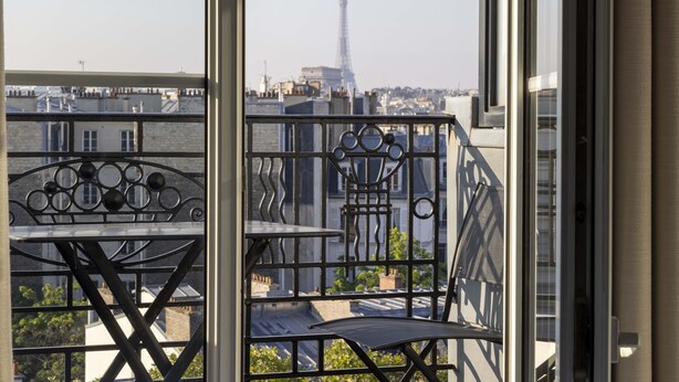 A balcony with a metal railing and a table, overlooking a cityscape with buildings and the Eiffel Tower in the distance. - Hôtel de Banville (Paris)
