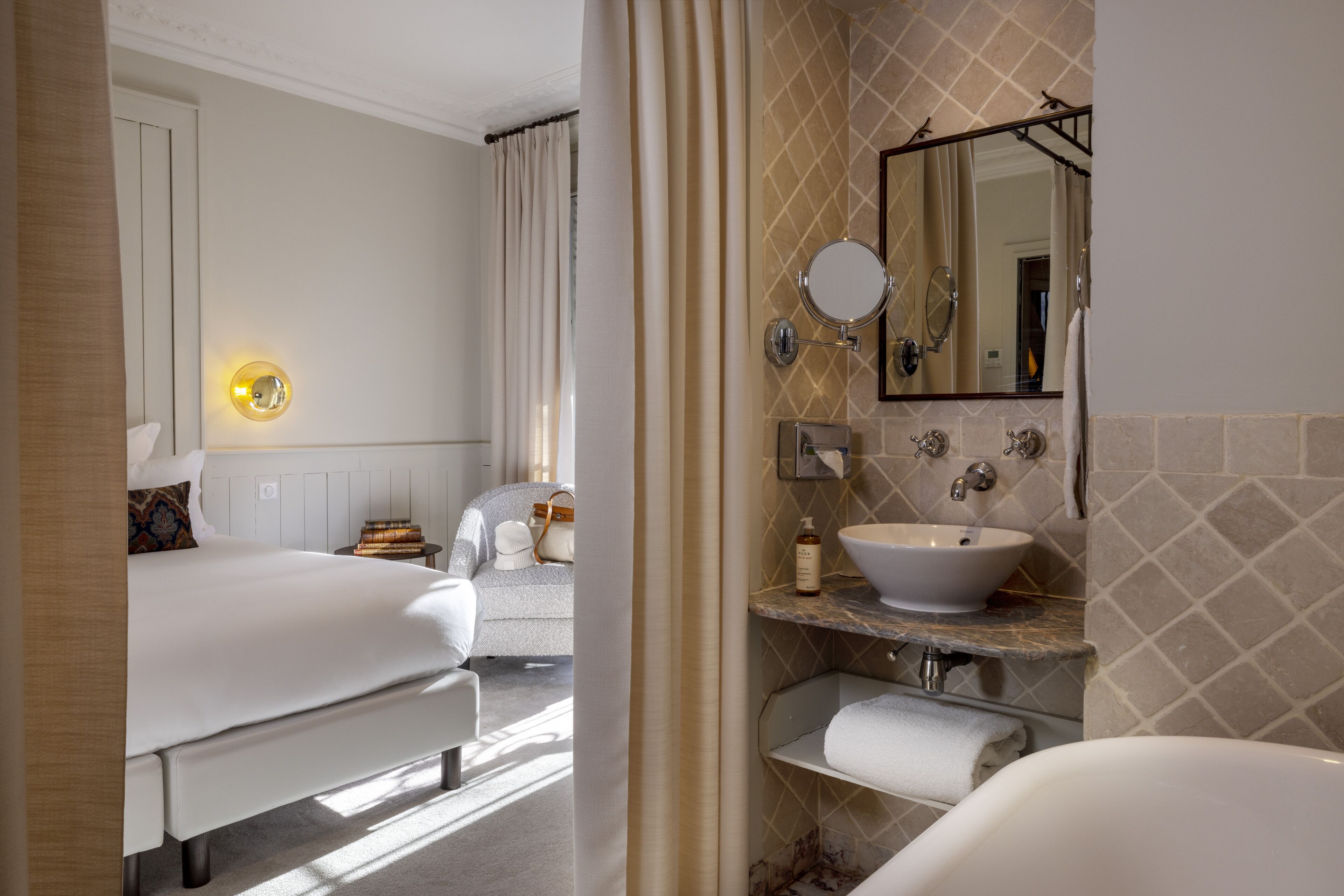 Una habitación de hotel con cama, baño con lavabo y espejo, y mesita de noche con lámpara.
