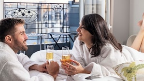 A couple in a hotel room, sitting on a bed with drinks, overlooking a balcony with a railing and chairs. - Hôtel de Banville (Paris)