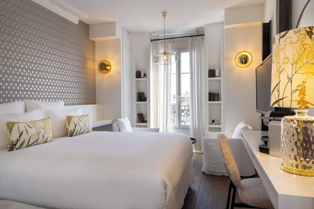 A hotel room with a large bed, a desk, a chair, a TV, and a bookshelf.