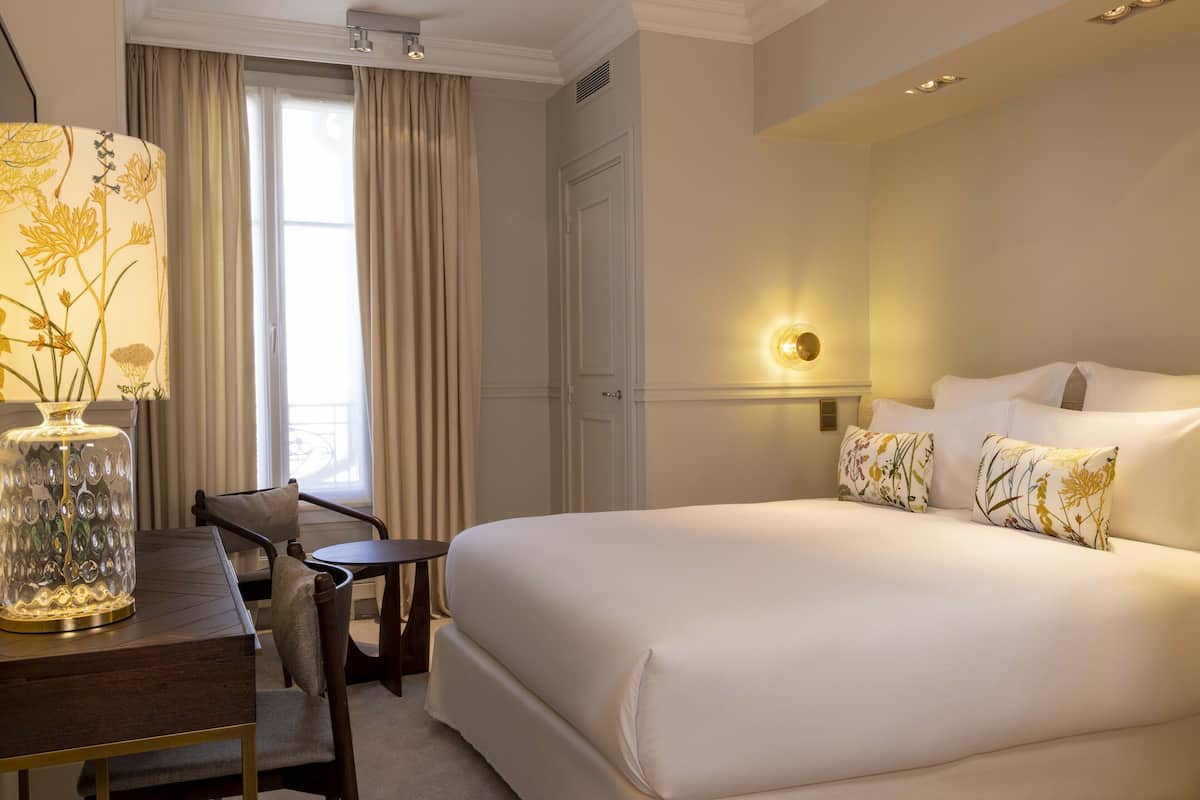 A hotel room with a large bed, a desk with a lamp, and a view of the outdoors through the window.