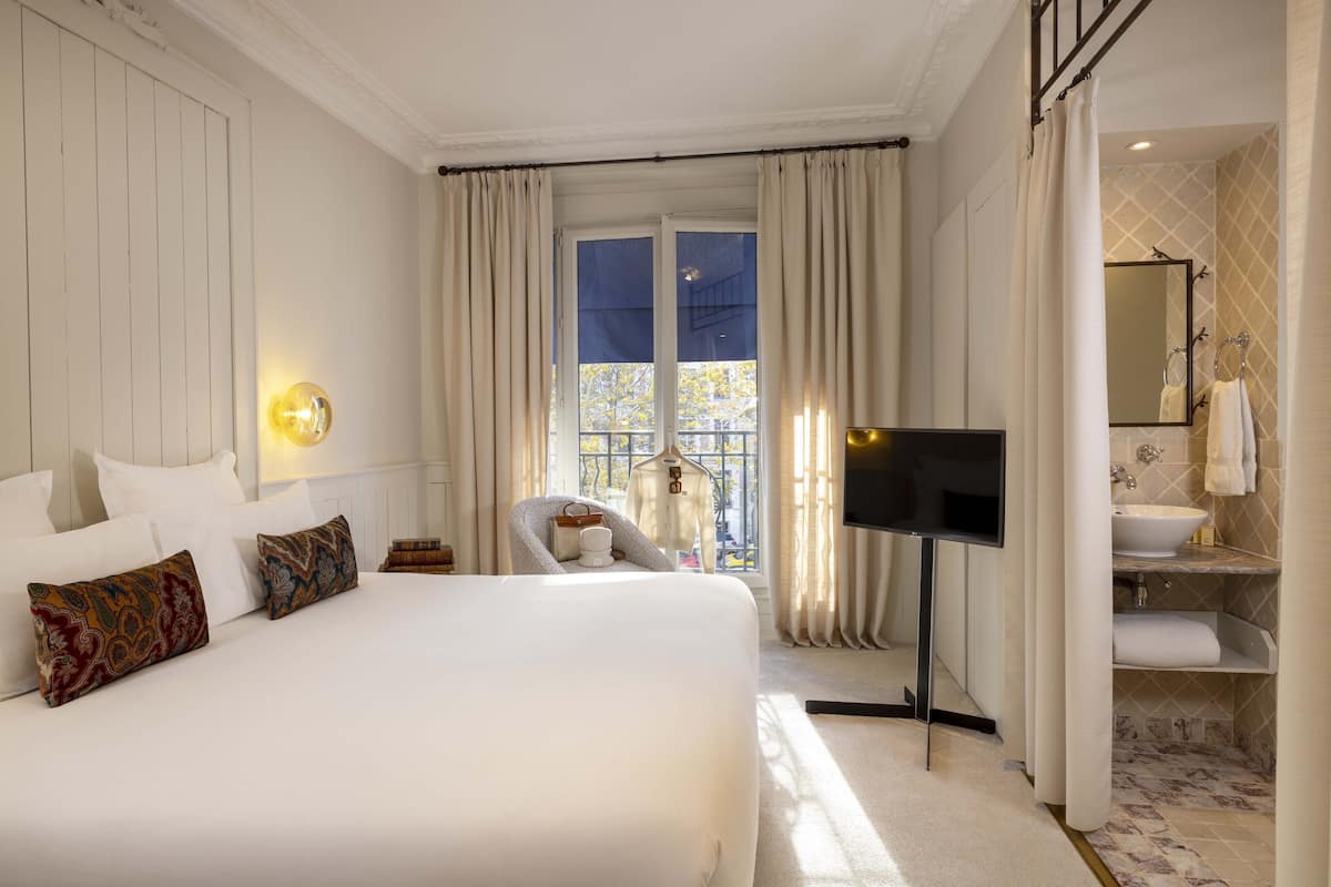 A hotel room with a large bed, a TV, a window with curtains, a chair, and a bathroom with a sink and mirror.