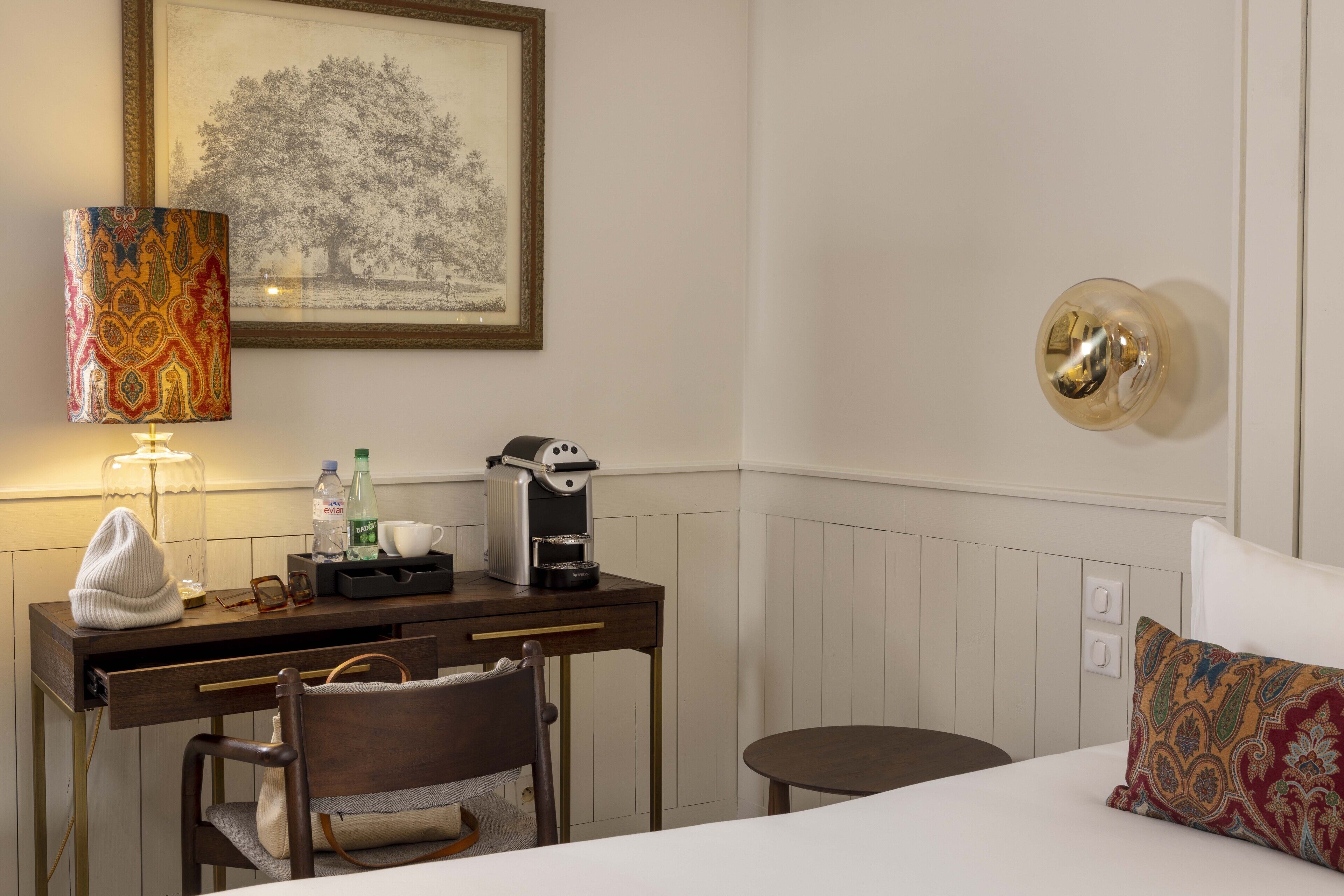 A hotel room with a bed, a desk with a lamp, a coffee maker, and a framed picture on the wall.