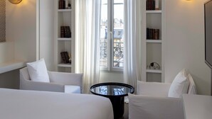 A hotel room with two white beds, a round black table, and a bookshelf with books and decorative items. - Hôtel de Banville (Paris)