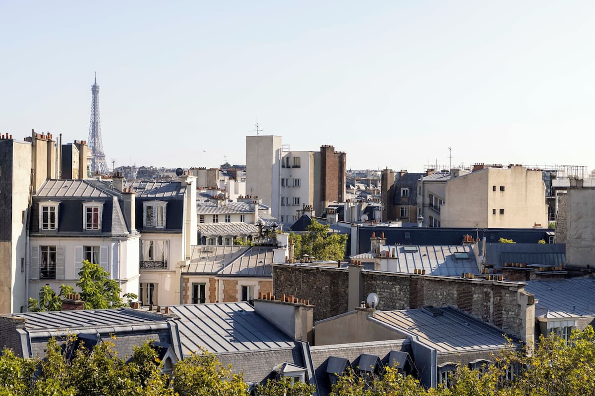 A cityscape with a prominent tower in the distance, likely the Eiffel Tower, surrounded by a mix of residential buildings with varying roof styles and some greenery.