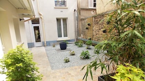 A courtyard with a gravel path, potted plants, and a building with a balcony. - Hotel Excelsior Batignolles (Paris)
