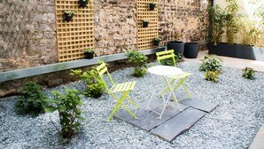 A patio with a stone wall, a wooden trellis with potted plants, a yellow chair, a white table, and a small garden area. - Hotel Excelsior Batignolles (Paris)