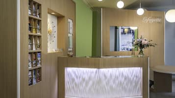 Un mostrador de recepción con detalles de madera, una pared verde y un espejo encima. Hay luces colgantes y un florero con flores sobre el mostrador.