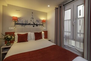 Una camera d'albergo con un letto grande, due abat-jour, un affresco raffigurante Parigi, una finestra con tende e un comodino con un vaso di fiori.