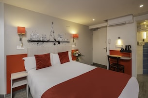 A hotel room with a bed, a desk, a chair, a wall-mounted air conditioner, and a wall decoration depicting Paris landmarks.