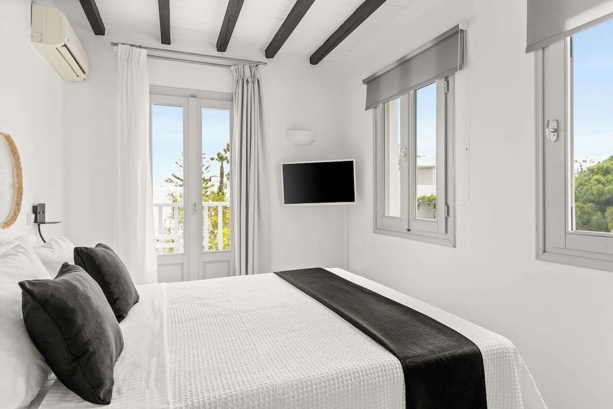 Habitación de hotel con cama, paredes blancas, televisión de pantalla plana y balcón con cortinas.