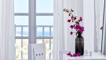 A white bedside table with a vase of pink flowers and a framed picture leaning against a window with white curtains.