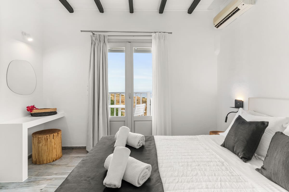 Un dormitorio moderno con una cama, un taburete de madera, una mesita con un florero y vistas al exterior a través de una ventana.