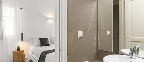 Un dormitorio moderno con cama, mesita de noche y velador. El baño cuenta con un lavamanos grande, espejo y perchero.