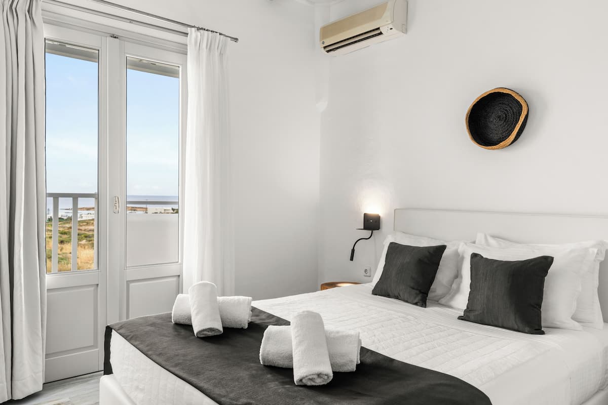 Habitación de hotel con cama, cortinas blancas, vista al mar y aire acondicionado instalado en la pared.