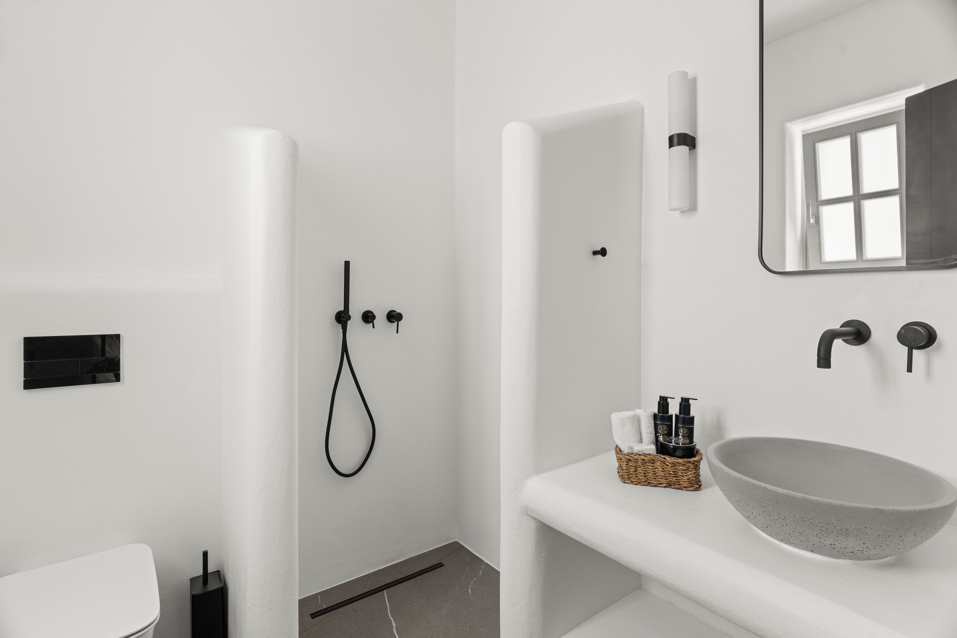 Une salle de bain moderne avec des toilettes blanches, un grand lavabo sur pied, une douche avec un pommeau de douche à main et un miroir avec une fenêtre au-dessus.