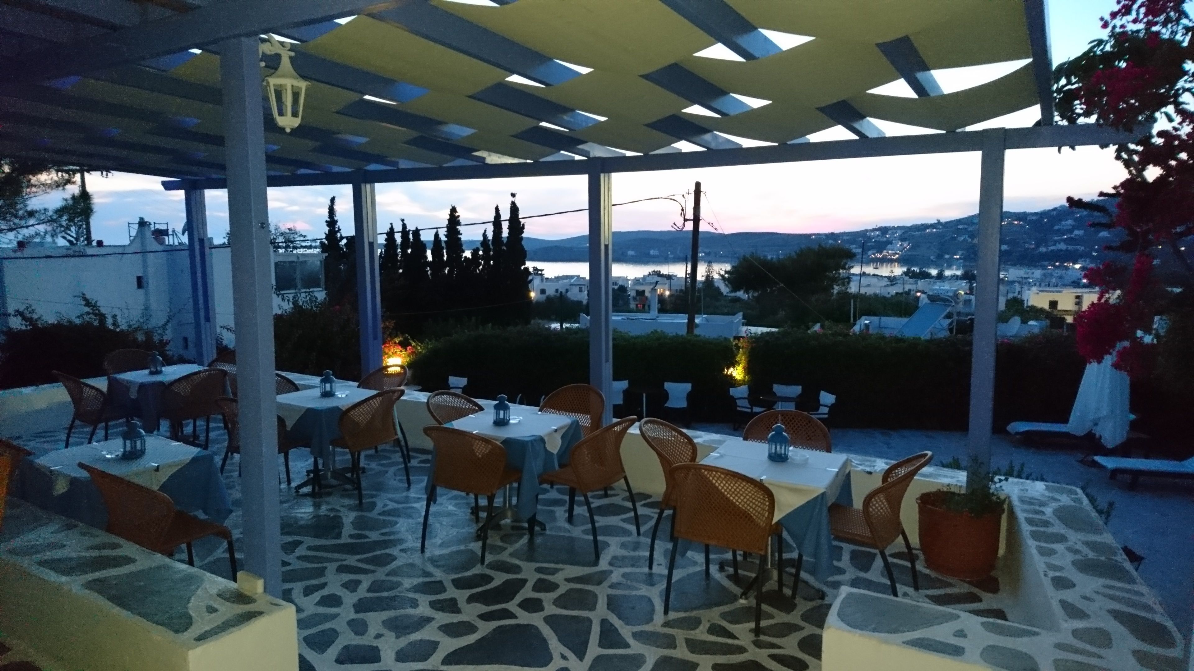 Ein Essbereich im Freien mit Tischen und Stühlen, einer Pergola und Blick auf eine Stadt und Hügel im Hintergrund.