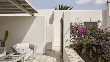 A sunlit patio with a white wall, a stone wall with a climbing plant, a wicker sofa, and a glass table with a bowl of fruit.