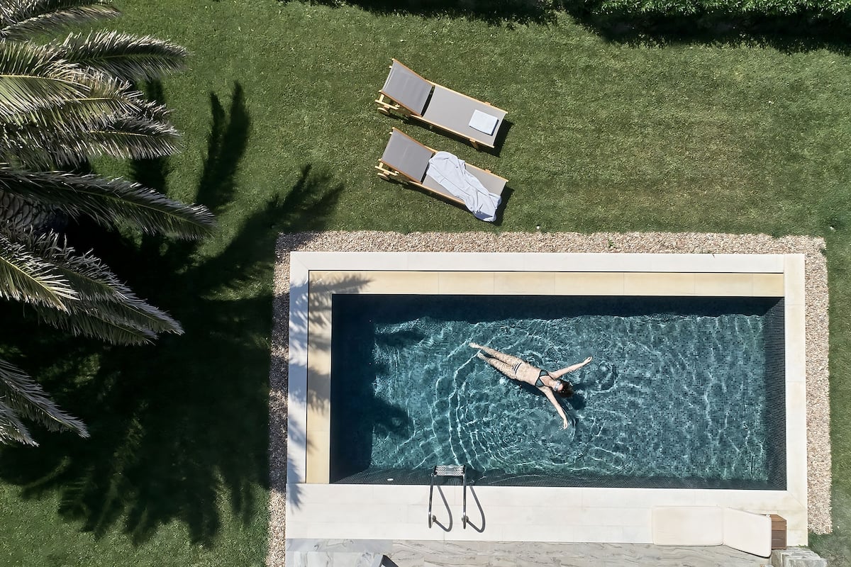 Vue aérienne d’une piscine où une personne nage, entourée d’une pelouse bien entretenue et de deux chaises longues.